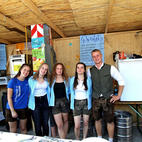 Fünf Frauen und ein Mann stehen in einem rustikalen Raum mit Holzwänden. Sie lächeln für ein Foto. Hinter ihnen befindet sich ein Speiseplan und ein Kühlschrank.
