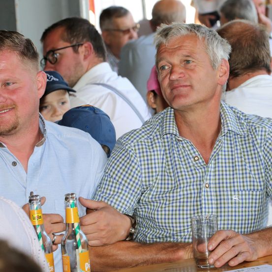 Zwei Männer sitzen an einem Tisch mit Getränken vor ihnen. Der Mann auf der linken Seite hat eine Bierflasche mit einem grünen Strohhalm, während der Mann auf der rechten Seite ein Glas hält. Sie sind von anderen Menschen umgeben.