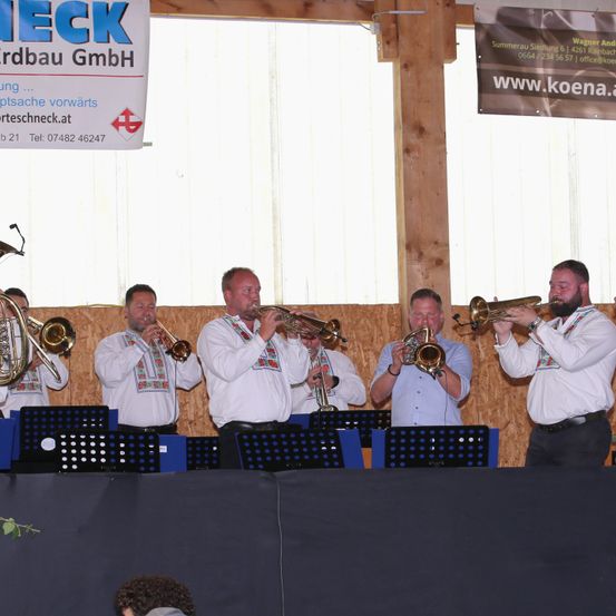 Eine Gruppe von Musikern in traditioneller Kleidung spielt auf der Bühne und musiziert mit Trompeten und Posaunen. Hinter ihnen befinden sich Banner mit Logos und Text.