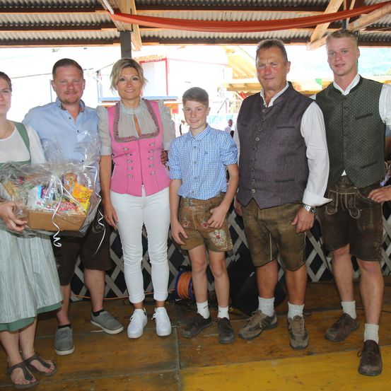 Eine Gruppe von Erwachsenen und Kindern in traditioneller Kleidung steht zusammen. Eine Frau hält einen Korb mit Geschenken. Die Umgebung scheint ein Ereignis oder eine Feier zu sein.