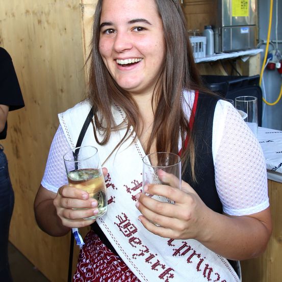 Eine lächelnde Frau in einem Festivalband hält zwei Gläser Bier. Sie steht neben einer Holzwand.