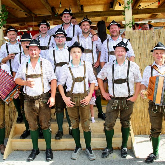 Eine Gruppe von Männern in traditioneller bayerischer Kleidung posiert für ein Foto vor einer Holzstruktur. Sie tragen Hosenträger, Hüte und halten Musikinstrumente.