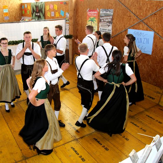 Eine Gruppe von Menschen in traditioneller Kleidung tanzt gemeinsam auf einem Holzboden. An der Wand befinden sich Plakate mit einer Veranstaltungsankündigung.