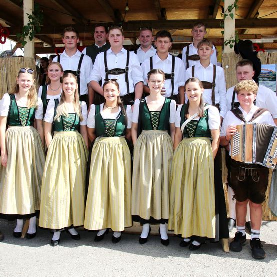 Eine Gruppe junger Menschen, gekleidet in traditioneller deutscher Tracht, posiert für ein Foto. Die Mädchen tragen grüne und gelbe Kleider mit weißen Schürzen. Die Jungen tragen weiße Hemden und Hosenträger. Ein Junge hält ein Akkordeon.