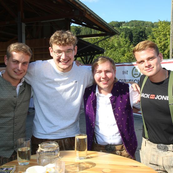 Vier Menschen lächeln und posieren für ein Foto im Freien, vor ihnen steht ein Tisch mit Getränken und eine malerische Kulisse.