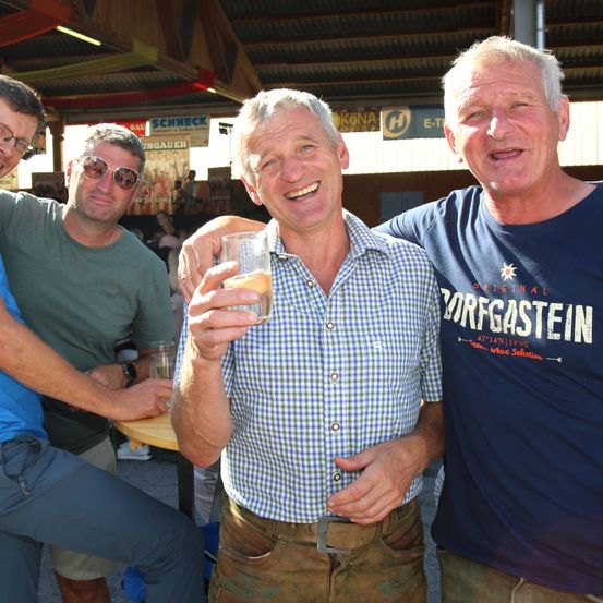 Drei Männer lächeln und posieren für ein Foto bei einer Outdoor-Veranstaltung, einer hält ein Getränk, ein anderer trägt ein grünes Shirt und der letzte ein blaues Shirt mit Text.