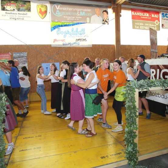 Eine Gruppe von Menschen tanzt auf einer Bühne bei einem Festival, umgeben von Holzwänden und Bannern. Einige tragen Freizeitkleidung, andere traditionelle Kleidung. Ein Mann in einem gestreiften Hemd steht in der Nähe eines Tisches.