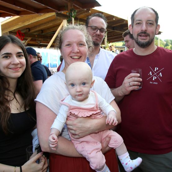 Ein Familienfoto zeigt eine Frau, die ein Baby mit lächelndem Gesicht hält, während ein Mann neben ihr ein Glas hält. Hinter ihnen schauen eine weitere Frau und ein Mann mit Brille zur Kamera. Im Hintergrund befindet sich eine Holzstruktur und ein Banner.