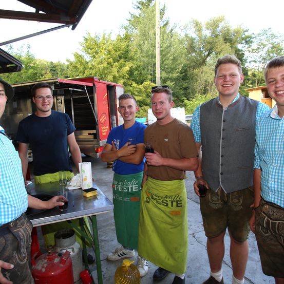 Fünf junge Männer stehen im Freien in der Nähe eines Essenswagens. Ein Mann hält ein Glas. Die Männer tragen Freizeitkleidung mit Schürzen, einer mit einem blauen Shirt, und andere in verschiedenen farbigen Shirts. Der Hintergrund zeigt Grünpflanzen und eine Holzstruktur.