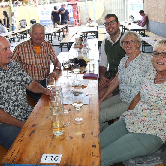 Fünf Personen sitzen an einem Tisch mit Weinläsern und Tellern. Sie lächeln und scheinen für ein Foto zu posieren. Es gibt andere Tische und Bänke in der Umgebung.