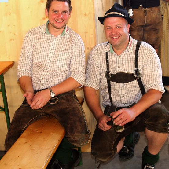 Zwei Männer in traditioneller bayerischer Kleidung sitzen auf einer Bank. Einer hält ein Bierglas, während der andere in die Kamera lächelt. Dahinter ist eine Holzwand.