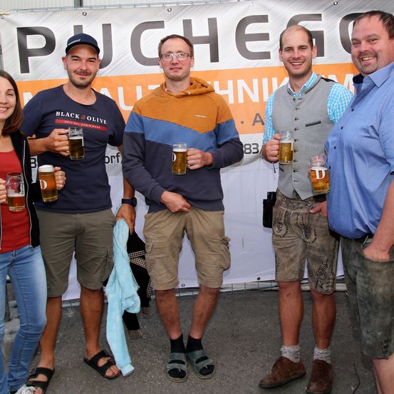 Fünf Personen stehen zusammen und halten Gläser mit Bier vor einem Banner, auf dem 'PUCHEGG' steht. Drei Männer tragen Shorts, einer trägt eine Weste. Eine Frau steht links.