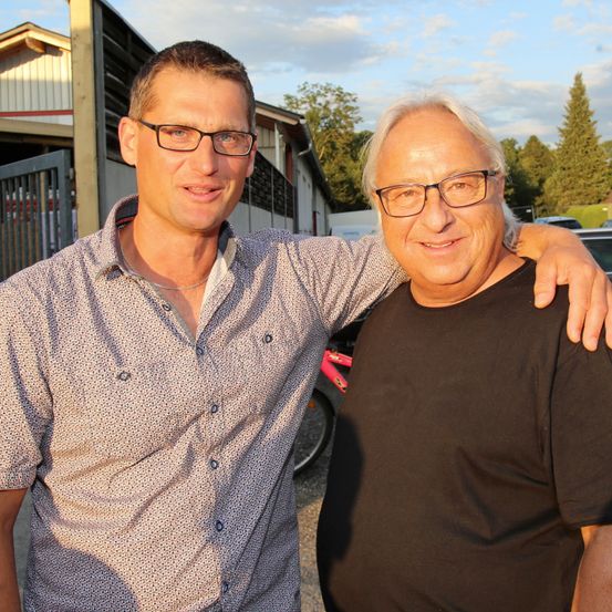 Zwei Männer stehen nebeneinander, einer mit der Hand auf der Schulter des anderen. Sie lächeln, tragen beide Brillen. Hinter ihnen steht ein Gebäude, und ein Auto ist in der Nähe geparkt.