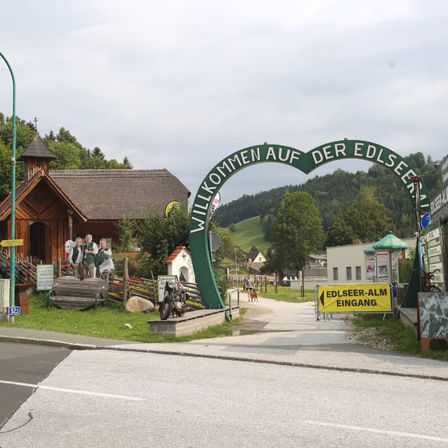 Eine Straße, die zu einem malerischen Dorf führt, mit einem grünen Bogen und einer Gruppe von Menschen in traditioneller Kleidung. Ein Schild weist auf den Eingang zum Edlsee Alm hin.