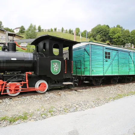 Eine schwarze Lokomotive mit grünem Logo und roten Rädern steht auf einem Gleis mit Kies und Gras. Ein grüner Anhänger ist angeschlossen.