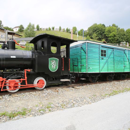 Eine schwarze Lokomotive mit grünem Logo und roten Rädern steht auf einem Gleis mit Kies und Gras. Ein grüner Anhänger ist angeschlossen.