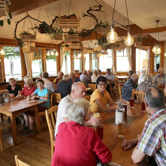 Eine Gruppe älterer Menschen sitzt an Tischen in einem Restaurant. Sie lächeln und scheinen eine gute Zeit zu haben. Die Tische sind mit Getränken und Essen gefüllt.