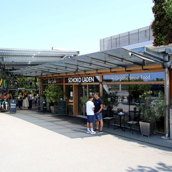 Zwei Männer stehen vor einem Café mit dem Namen Schoko Laden, was Schokoladenladen bedeutet. Das Café hat eine Metallüberdachung, Glaswände und Topfpflanzen. Menschen gehen auf dem Bürgersteig und ein Gebäude mit Solarpanelen ist im Hintergrund zu sehen.