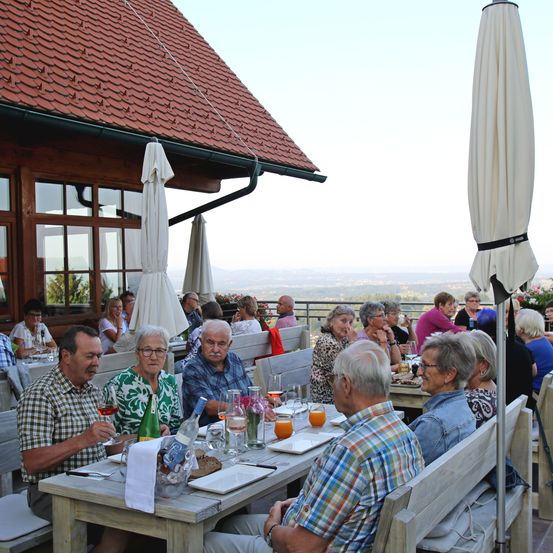 Eine Gruppe älterer Menschen isst im Freien und sitzt um Tische herum, mit Getränken und Essen. Sie sind unter Schirmen, mit Blick auf die Berge im Hintergrund.