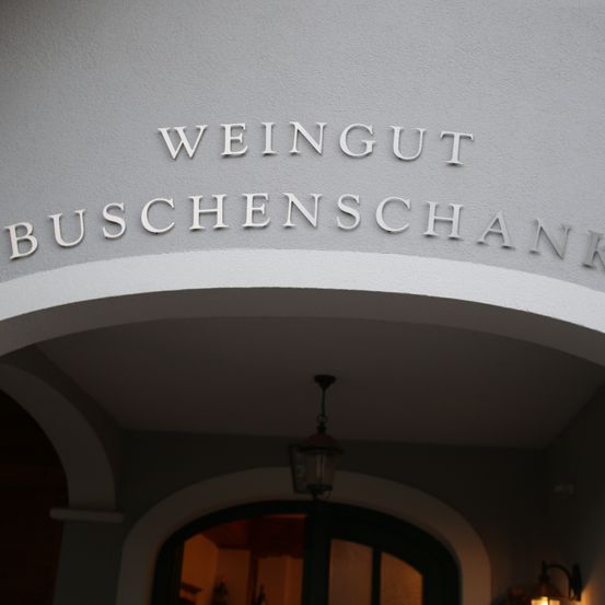 Der Eingang einer Buschenschank, einer Weinschenke, ist zu sehen. Der Name ist weiß auf einer grauen Wand geschrieben. Eine Lampe hängt von der Decke.
