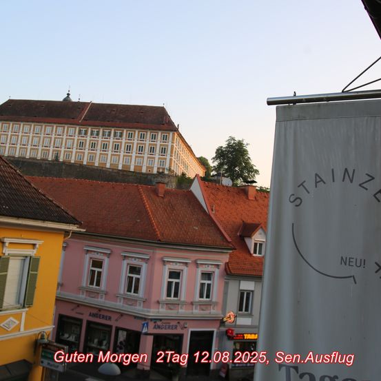 Ein Blick auf die Straße mit Gebäuden im Hintergrund, eines mit einem Schild, das 'Guten Morgen 2Tag 12.08.2025, Sen.Ausflug' steht. Das Gebäude hat ein Banner mit dem Wort 'STAINZ' und einem lächelnden Gesicht.
