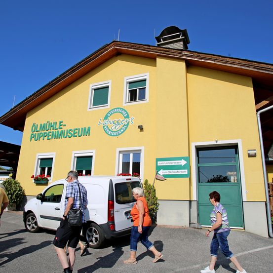 Mehrere Menschen gehen an einem weißen Van vorbei, der vor einem gelben Gebäude mit grünen Fenstern steht, das mit 'Olmuhle-Puppenmuseum' beschriftet ist. Eine Frau in einem orangefarbenen Shirt und ein Mann in einem karierten Hemd sind darunter.