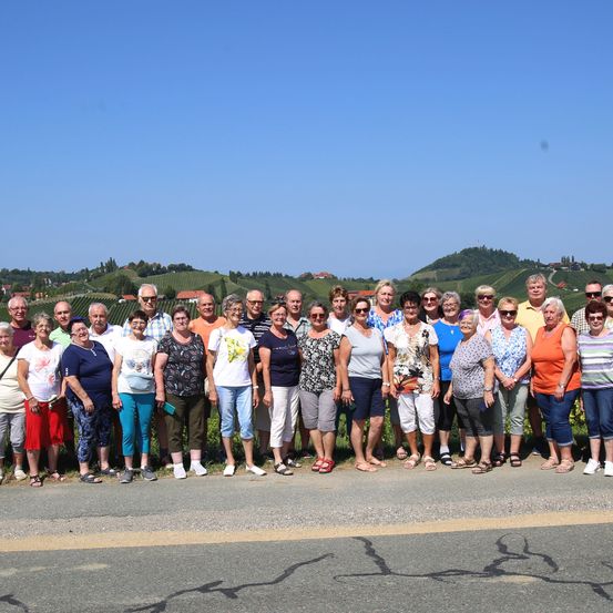 Eine Gruppe von Menschen, die auf der Straße stehen, Sonnenbrillen und Freizeitkleidung tragen. Dahinter eine Landschaft mit Bergen und Grün.