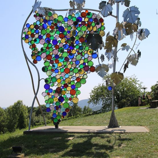 Eine große, farbenfrohe Glas-Skulptur in Form einer Traube steht auf einem Grashügel unter klarem Himmel. Sie besteht aus vielen kleinen Glaskreisen in verschiedenen Farben.