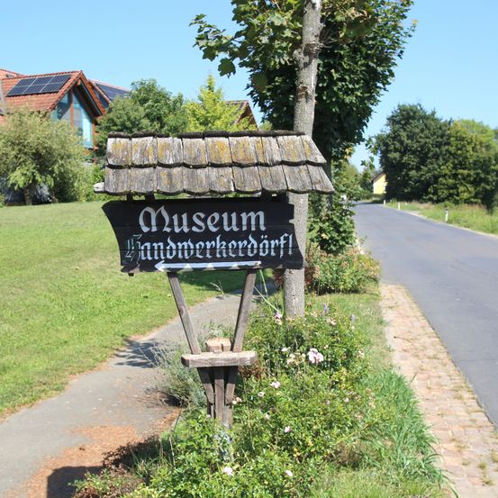 Ein Wegweiser zum örtlichen Museum steht am Straßenrand, mit einem Baum, einem Haus mit Solarpanelen und einer Rasenfläche im Hintergrund.
