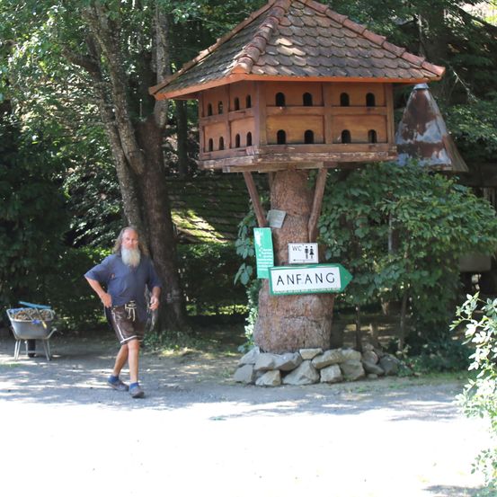 Ein bärtiger Mann geht in der Nähe eines großen Vogelhauses, das an einem Baum befestigt ist, mit einem Schild, das auf 'Anfang' zeigt, und einem Toilettenschild in der Nähe.