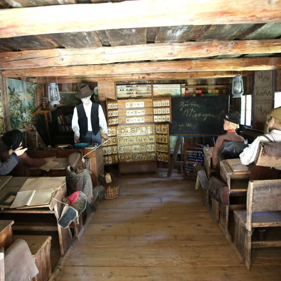 Ein hölzernes Klassenzimmer mit Schülern und einem Lehrer. Der Lehrer steht vor einer Tafel mit deutschem Text. Holzbänke und Tische sind in Reihen angeordnet. Eine Laterne hängt an der Wand.