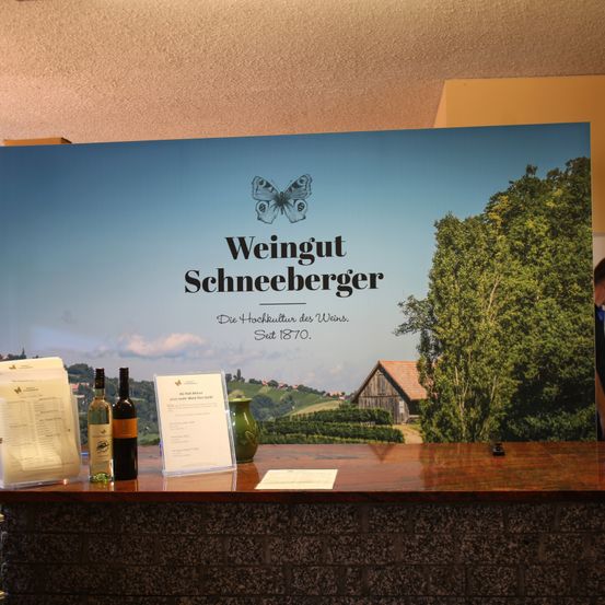 Ein großes Poster für Weingut Schneeberger zeigt einen malerischen Weinberg. Flaschen Wein, eine Speisekarte und eine grüne Vase stehen auf einem Holztresen.