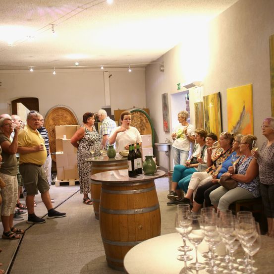 Eine Gruppe von Menschen versammelt sich um Holzfässer in einem Raum und probiert Wein. Einige stehen, andere sitzen. Flaschen und Weingläser stehen auf dem Tisch. Kunstwerke hängen an den Wänden.
