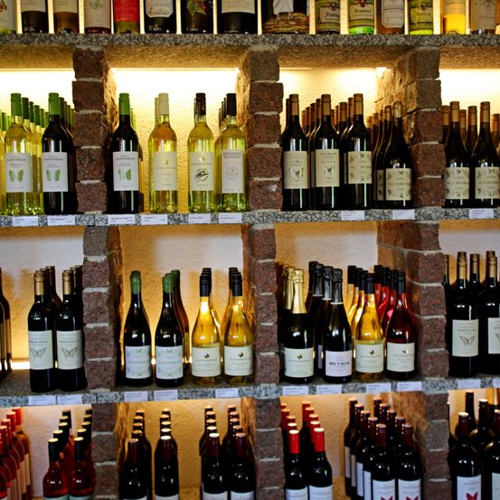 Ein Weinladen mit mehreren Regalen voller Weinflaschen, einschließlich Rot-, Weiß- und Schaumweinen, präsentiert in einer steingemauerten Umgebung. Jede Flasche hat ein Etikett mit einem Schmetterlingssymbol.