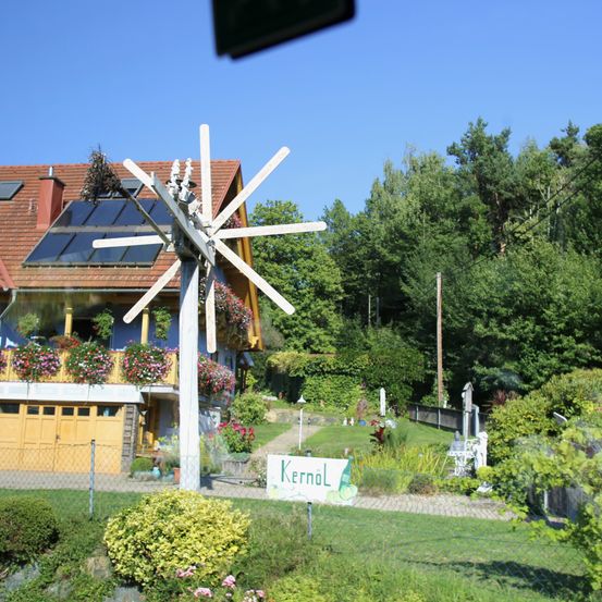 Ein Haus mit Solarpanelen auf dem Dach und einer Windmühle im Garten. Das Haus ist von einem Zaun umgeben, und ein Schild mit der Aufschrift 'Kernol' ist zu sehen.