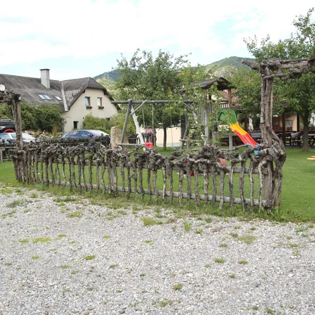 Ein Weg führt zu einem Haus mit Schaukel und Rutsche, umgeben von einem rustikalen Holzzaun. Bäume und Büsche sind sichtbar, mit Bergen im Hintergrund.