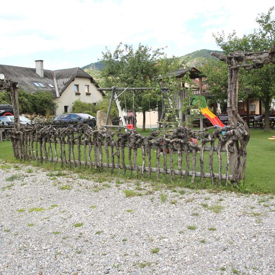 Ein Weg führt zu einem Haus mit Schaukel und Rutsche, umgeben von einem rustikalen Holzzaun. Bäume und Büsche sind sichtbar, mit Bergen im Hintergrund.