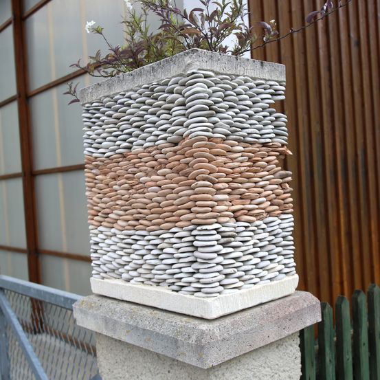 Ein quadratischer Pflanzbehälter aus Beton, verziert mit einer Vielzahl farbiger Kieselsteine, die in horizontalen Schichten angeordnet sind. Der Pflanzbehälter steht auf einem Betonsockel mit einem Metallzaun dahinter.