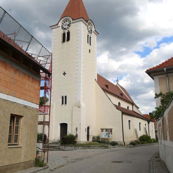 Eine weiße Kirche mit einem roten Dach und einem hohen Turm steht in der Mitte einer Straße. Der Turm hat auf beiden Seiten eine Uhr. Vor der Kirche befindet sich ein kleiner Garten.