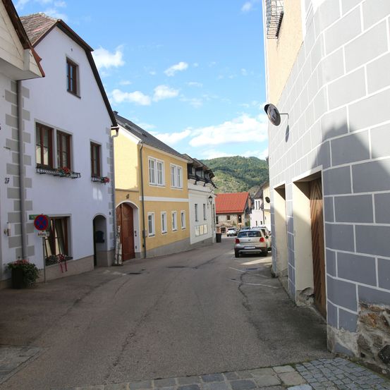 Eine schmale Straße mit zwei geparkten Autos. Gebäude mit Balkonen und Fenstern säumen beide Seiten. Ein Berg ist in der Ferne sichtbar.