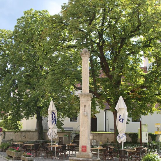 Ein Denkmal steht in einem von Bäumen umgebenen Innenhof. Tische und Stühle sind unter Schirmen aufgestellt. Ein Gebäude mit Treppen und Bänken befindet sich hinter dem Innenhof.