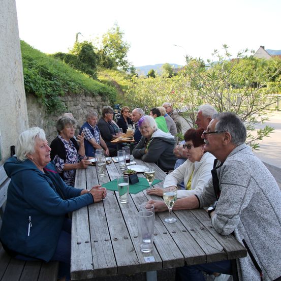 Eine Gruppe älterer Menschen sitzt um einen Holztisch herum, vielleicht bei einem Picknick. Sie alle lächeln und scheinen gut gelaunt zu sein.