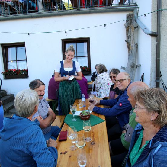 Ein Außenbereich zum Essen mit einer stehenden und lächelnden Frau, möglicherweise eine Kellnerin. Mehrere ältere Menschen sitzen um einen Holztisch herum und unterhalten sich möglicherweise. Auf dem Tisch stehen Gläser, eine Topfpflanze und ein Notizbuch.
