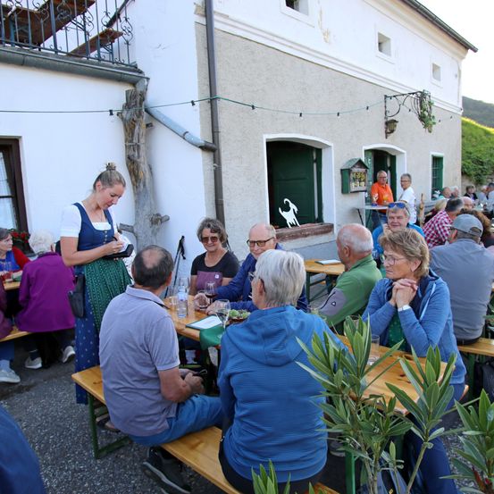 Eine Gruppe von Menschen sitzt um Tische herum außerhalb eines Gebäudes, eine Frau in traditioneller Kleidung serviert ihnen. Die Tische haben Pflanzen, Weingläser und Essen. Eine Frau steht in der Nähe des Eingangs und ein Mann schaut sich etwas an.