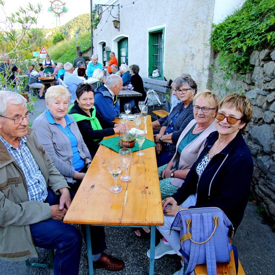 Eine Gruppe älterer Menschen sitzt um einen langen Holztisch im Freien und lächelt. Gläser und Pflanzen stehen auf dem Tisch. Dahinter befindet sich eine Steinmauer mit Grünpflanzen.