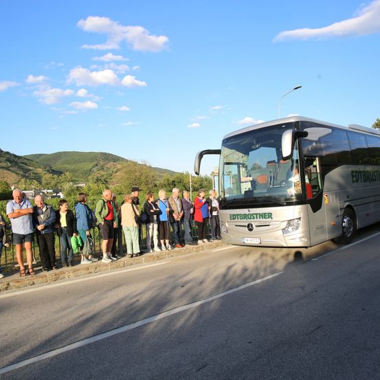 Eine Gruppe von Menschen steht am Straßenrand und beobachtet einen silbernen Bus mit dem Namen Edtbrustner. Der Bus ist geparkt und zeigt mit der Front auf die Gruppe. Im Hintergrund befindet sich ein Hügel mit Bäumen und ein blauer Himmel mit Wolken.