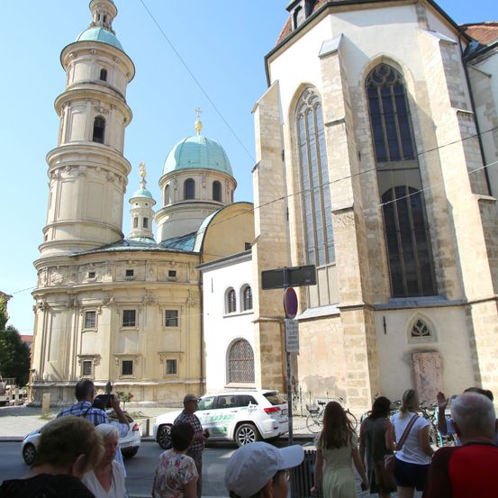 Menschen versammeln sich vor einer historischen Kirche mit gotischer Architektur. Die Kirche hat einen hohen Turm, eine Kuppel und große Spitzbogenfenster. In der Nähe ist ein weißer SUV geparkt, und ein Mann macht Fotos.