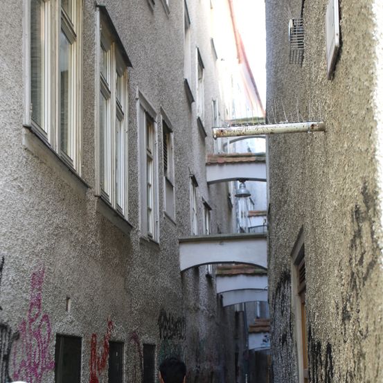 Ein schmaler Gang zwischen zwei alten Gebäuden mit Graffiti an den Wänden und mehreren Fenstern. Rechts ist eine Terrasse mit einer Lampe zu sehen.