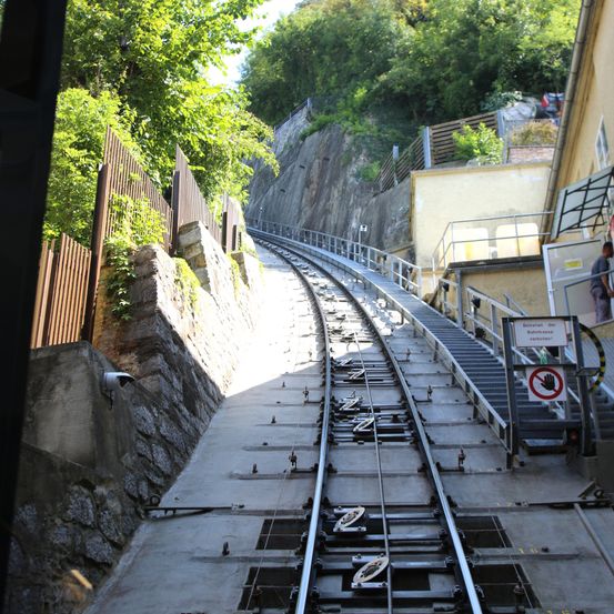 Ein Blick aus einem Zug auf Gleise an einem steilen Hang, mit einer Steinmauer links und einem Gebäude mit Treppen rechts.