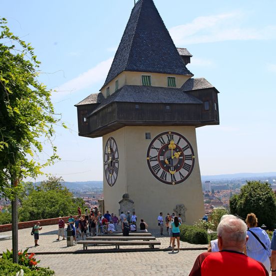 Ein großer Uhrturm mit einem dreieckigen Dach und einer sichtbaren Uhr, umgeben von einer Menschenmenge. Der Turm befindet sich in einem Park mit Blick auf die Stadt in der Ferne.
